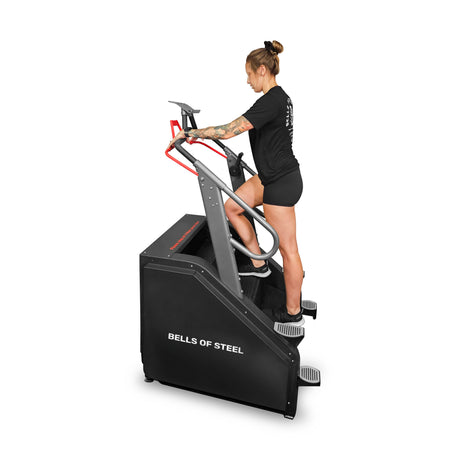 A tattooed person uses a Bells of Steel Sisyphean Stepper, donning a black shirt and shorts in their home gym. The sleek cardio machine in black with red handles aids their upward strides effectively.