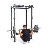 Wearing a black shirt and shorts, a person performs a squat with a barbell on their shoulders inside the Bells of Steel Manticore Four Post Power Rack Builder (3" x 3", 1" Holes). This home gym essential features adjustable safety bars and weight plate storage hooks for an efficient workout.