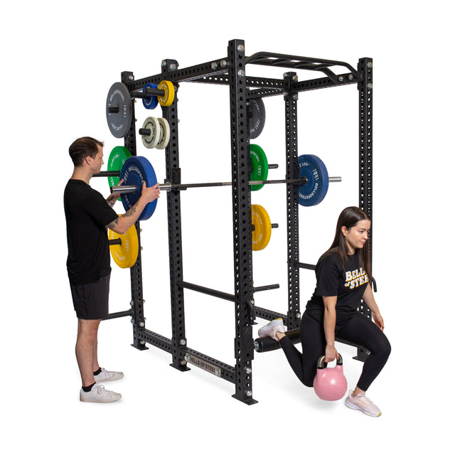 A man and a woman exercise near the Bells of Steel Manticore Six Post Power Rack, adjusting weights on a barbell with vibrant plates. The man handles the setup while the woman lunges with a pink kettlebell, both dressed in athletic wear.