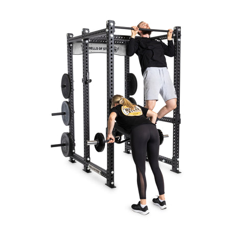 A man in a black shirt and grey shorts performs pull-ups on the Bells of Steel Manticore Six Post Power Rack (3" x 3", 1" Holes) while a woman in a black shirt and leggings deadlifts with a barbell, showcasing impressive strength within the gym's open ecosystem.