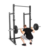 A man squats with a barbell on his shoulders, wearing a black shirt and gray shorts. He exercises on the Bells of Steel Manticore Half Rack Builder (3" x 3", 1" Holes) with modular components and a pull-up bar, isolated against a white background.