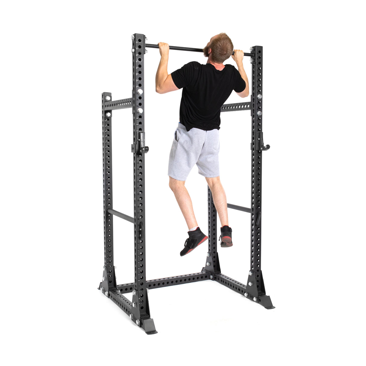 A person in a black t-shirt and gray shorts is doing a pull-up on the Manticore Half Rack Builder by Bells of Steel, displaying its modular features against a clean white background.