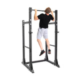 A person in a black t-shirt and gray shorts is doing a pull-up on the Manticore Half Rack Builder by Bells of Steel, displaying its modular features against a clean white background.