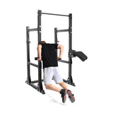 Wearing a black shirt and gray shorts, someone performs a dip exercise on the Manticore Half Rack Builder by Bells of Steel. Featuring adjustable bars and a pull-up bar at the top, this robust 3" x 3" rack supports a dip attachment, as their feet hover slightly off the ground.