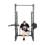 A man in a black t-shirt and shorts is kneeling in the Bells of Steel Manticore Half Rack Builder (3" x 3", 1" Holes) with modular components like a pull-up bar and dip handles. The open ecosystem is set up in a well-lit space.