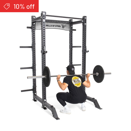 A person in black workout clothes squats with a barbell inside the Bells of Steel Manticore Collegiate Power Rack Builder (3" x 3", 1" Holes) at home. A red "10% off" label appears in the top left corner.