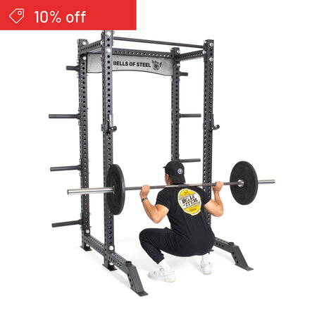 A person wearing black workout clothes squats with a loaded barbell inside a Bells of Steel Manticore Collegiate Power Rack Builder (3" x 3", 1" Holes). A red "10% off" banner appears in the top left corner. Ideal for any home gym.