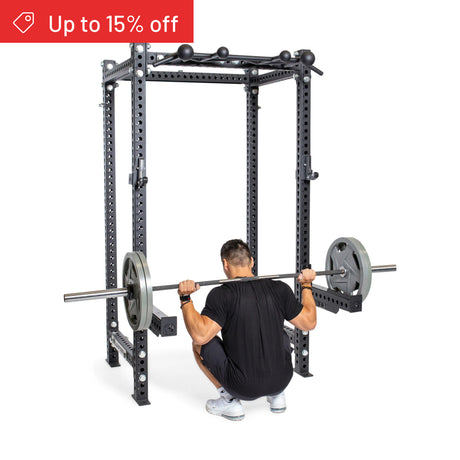 A man squats with a barbell inside the Bells of Steel Manticore Four Post Power Rack Builder (3" x 3", 1" Holes), featuring pull-up bars and safety rails. Ideal for home gyms. A red banner announces "Up to 15% off.