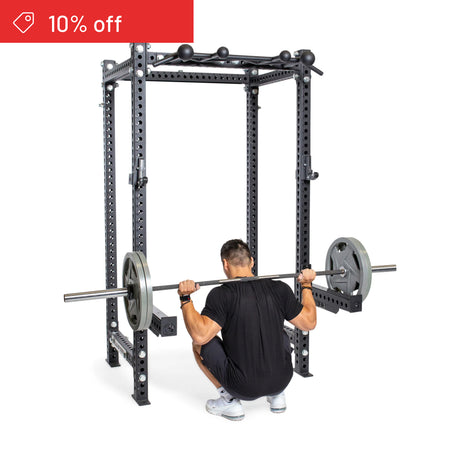 A man squats with a barbell inside the Bells of Steel Manticore Four Post Power Rack Builder (3" x 3", 1" Holes), viewed from behind. A red "10% off" tag appears—ideal for your home gym upgrade.