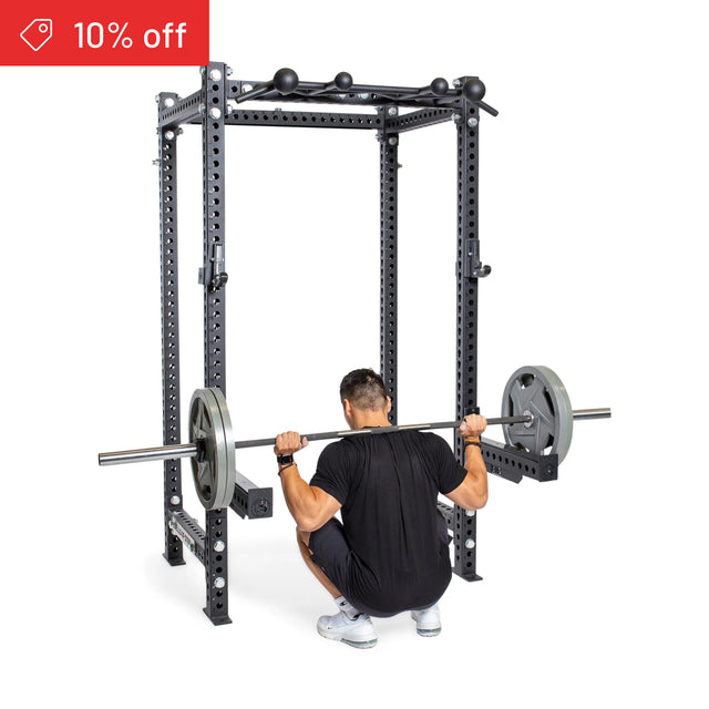A man squats with a barbell inside the Bells of Steel Manticore Four Post Power Rack Builder (3" x 3", 1" Holes) on a white background. A red "10% off" banner appears in the top left corner.