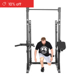 A man in a black t-shirt and shorts kneels inside the Bells of Steel Manticore Half Rack Builder (3" x 3", 1" holes). A "10% off" discount tag is shown in the top left, making it ideal for your home gym setup.