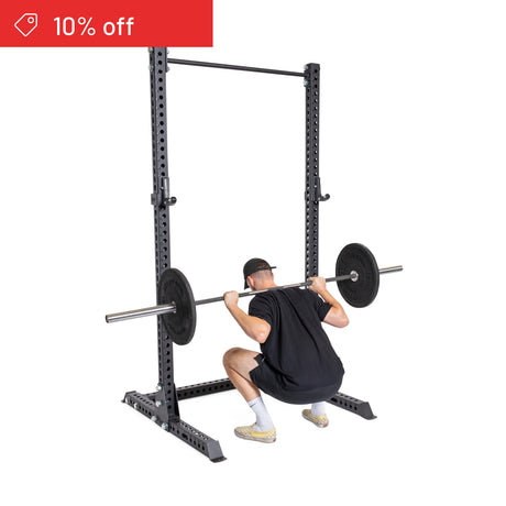 A person does a barbell squat on the Bells of Steel Manticore Squat Stand Builder (3" x 3", 1" holes), wearing a black shirt, shorts, cap, and white socks. A red "10% off" label appears on the white background—ideal for home gyms.