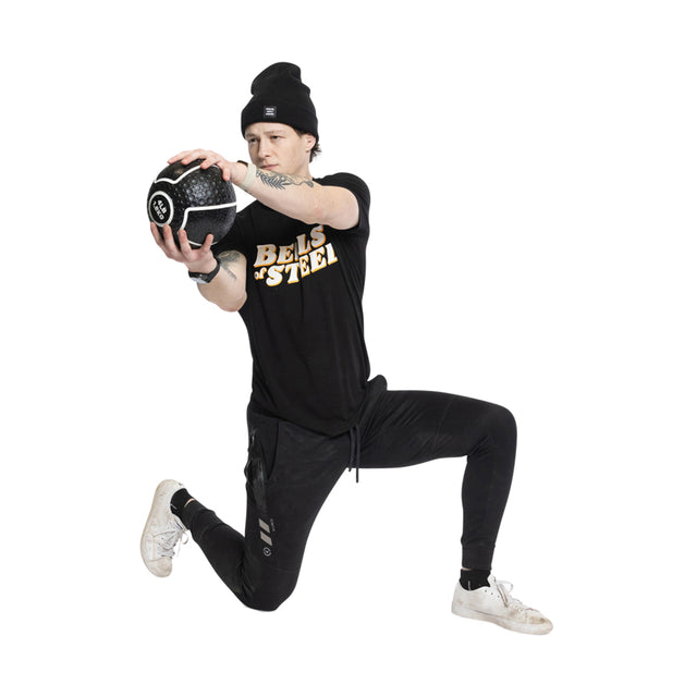 Kneeling on one knee, a person in a black beanie, T-shirt, and pants grips a Bells of Steel Mighty Grip Medicine Ball with focus and superior grip, showcasing their dedication during their multi-level workout.