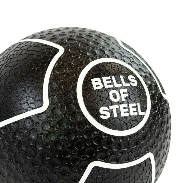 Close-up of a black Mighty Grip Medicine Ball by Bells of Steel shows its dimpled texture for superior grip. White curved lines and the brand name "BELLS OF STEEL" in white are centered, making it ideal for all fitness levels.