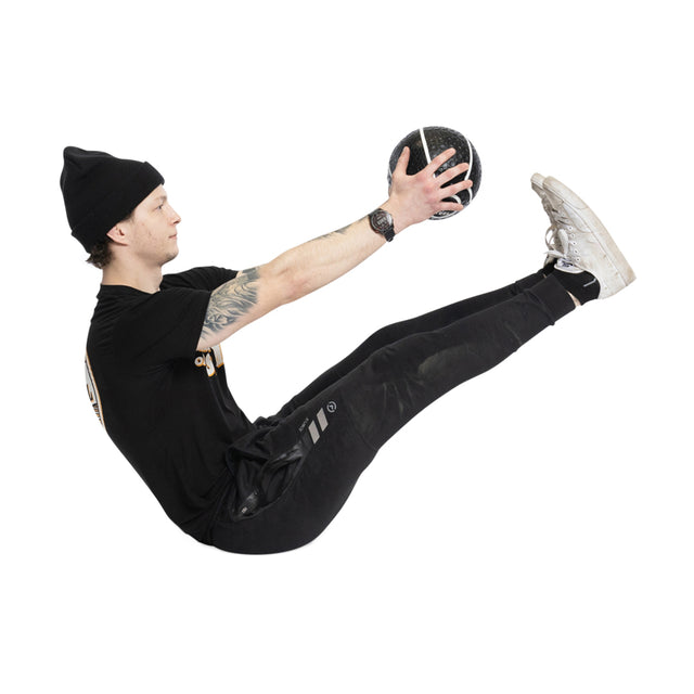 Wearing a black outfit and beanie, a person sits in a V-up position using a Bells of Steel Mighty Grip Medicine Ball. They balance impressively on their sitting bones with legs straight, feet raised off the ground, and arms extended while holding the ball.