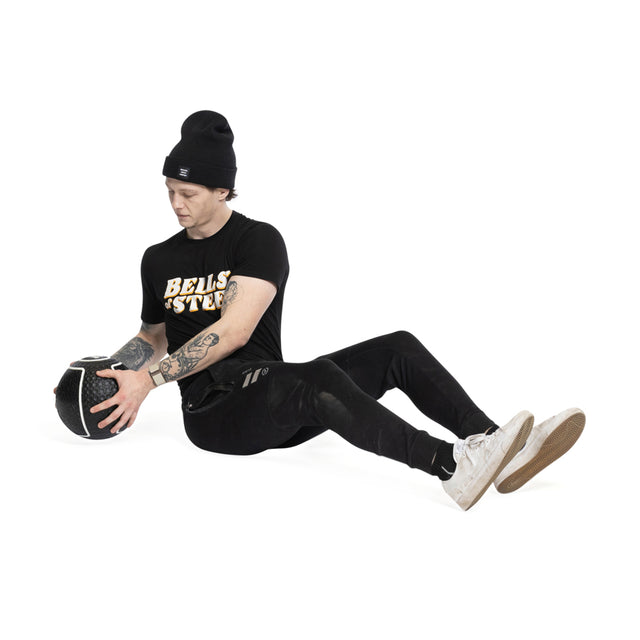 Dressed in a black beanie and t-shirt, the person sits on the floor with legs crossed, gripping a Mighty Grip Medicine Ball from Bells of Steel. The stark white background highlights their dedication to enhancing fitness using this top-quality grip essential.