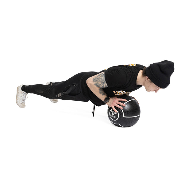 A person in a black outfit and beanie demonstrates impressive fitness by doing a plank with their hands on a Bells of Steel Mighty Grip Medicine Ball, labeled "Wall Ball 6 KG," against a white background.