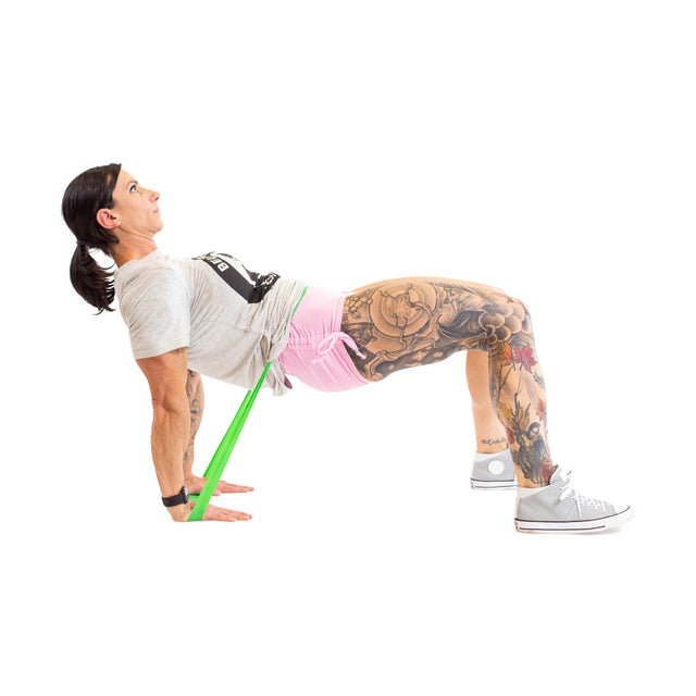 A person with tattooed legs performs a bridge exercise using Bells of Steel Mini Bands. They're wearing a gray T-shirt, pink shorts, and gray sneakers on a white backdrop, showcasing strength and fitness.