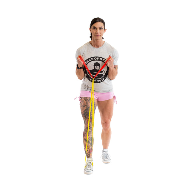 A person in a gray t-shirt and pink shorts demonstrates a gym routine with Bells of Steel Mini Bands. They stand with one foot forward on the band, pulling the handles upward, revealing tattoos on their left leg while wearing white sneakers.