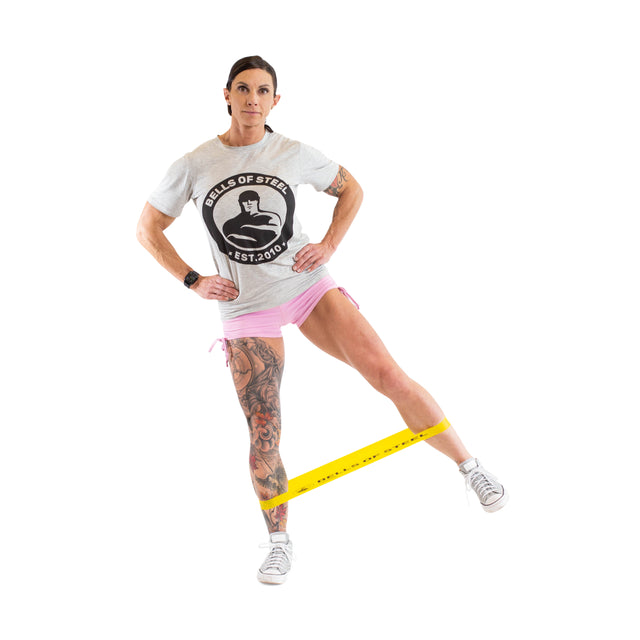 Wearing a gray T-shirt and pink shorts, a person uses Bells of Steel Mini Bands in their gym routine, performing lateral leg movements with a yellow band around their ankles. They balance on one leg and extend the other to the side for an effective workout.