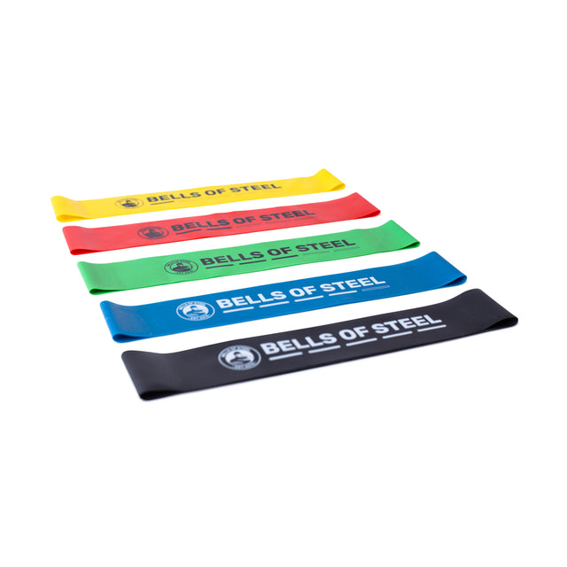 Five Bells of Steel Mini Bands in yellow, red, green, blue, and black are laid out in a row. Perfect for any gym routine, they feature the brand's logo.