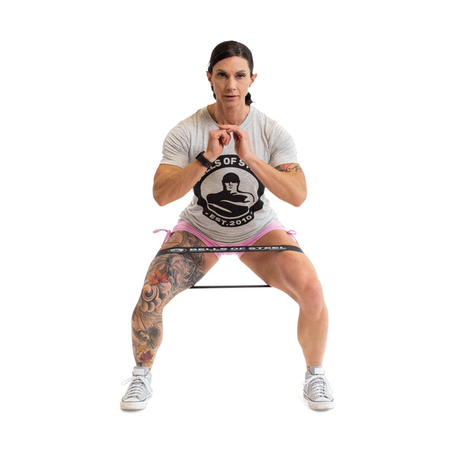 A person with short dark hair, wearing a light gray t-shirt and pink shorts, squats using Bells of Steel Mini Bands around their thighs. Their right leg sports a colorful tattoo.