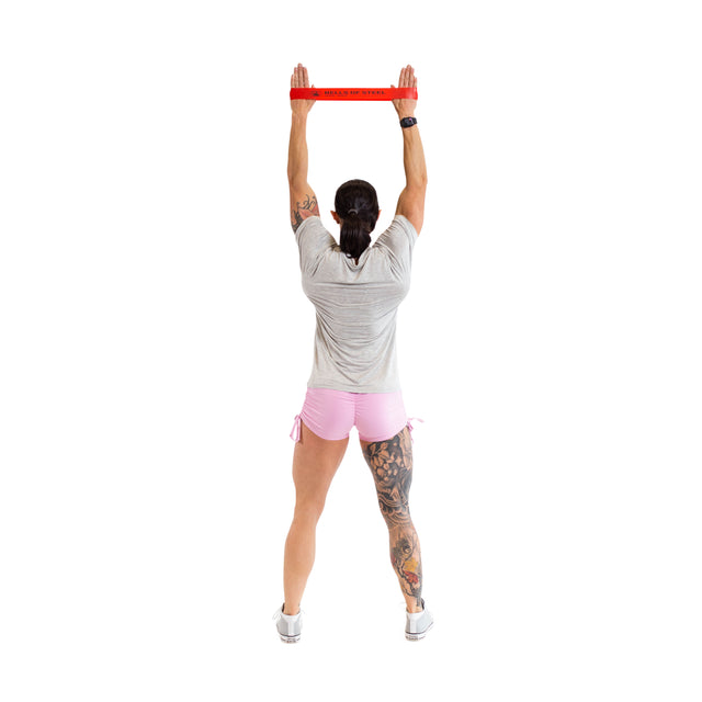 A person with tattoos on one leg confidently stretches a red Bells of Steel Mini Band above their head, engaged in a routine wearing a gray shirt and pink shorts against a white background.