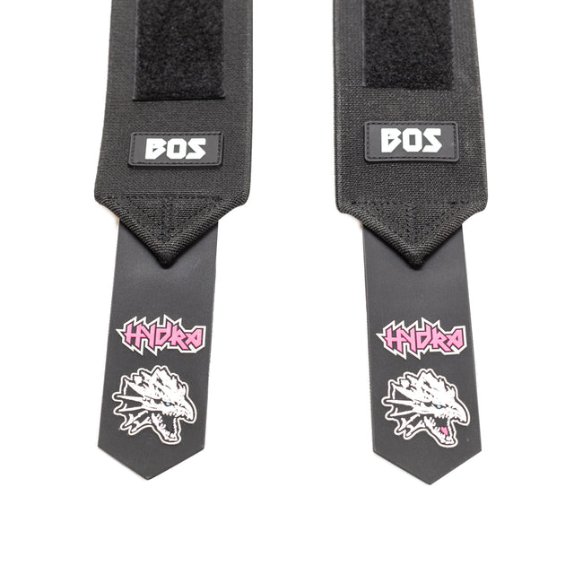 Two black Mighty Wrist Wraps by Bells of Steel with "BOS" labels and end tabs featuring a white and pink "HYDRA" logo above a dragon skull. These powerlifting essentials provide wrist support and stand out on a white background.