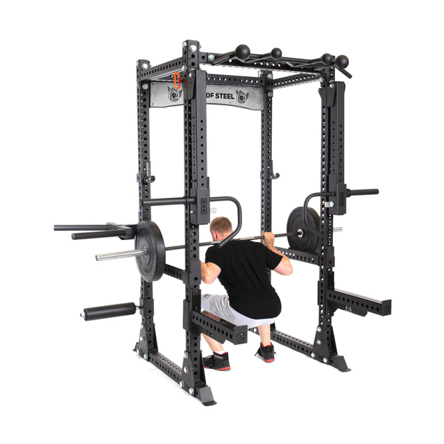 A person is squatting in a Bells of Steel Manticore Flat Foot Power Rack, with a barbell on their shoulders. This 1,000-pound capacity rack, equipped with modular weights and safety bars, supports them as they wear a black shirt and shorts.