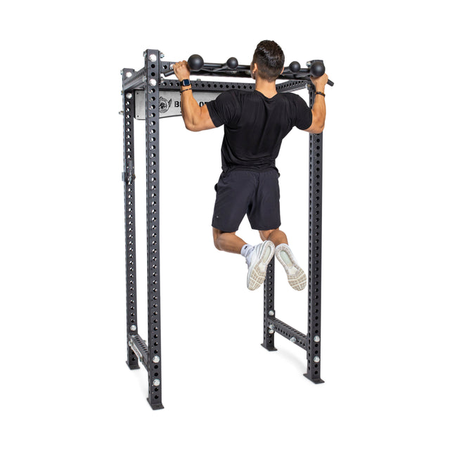 Someone in black athletic wear performs a pull-up on the Bells of Steel Globe Pull Up Bar, attached to a squat rack, building grip strength against a plain white background.
