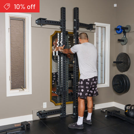 Someone adjusts the Bells of Steel Manticore Folding Half Rack Builder (3" x 3", 1" holes) in a home gym filled with weight plates and other gear. A red "10% off" sale banner appears at the top left of the image.