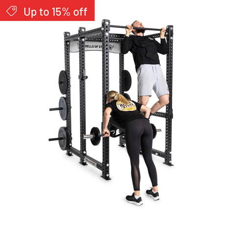 A man does pull-ups on the Bells of Steel Manticore Six Post Power Rack Builder (3"x3", 1" Holes) while a woman lifts a barbell—ideal for any home gym. A red banner in the top left corner reads "Up to 15% off.