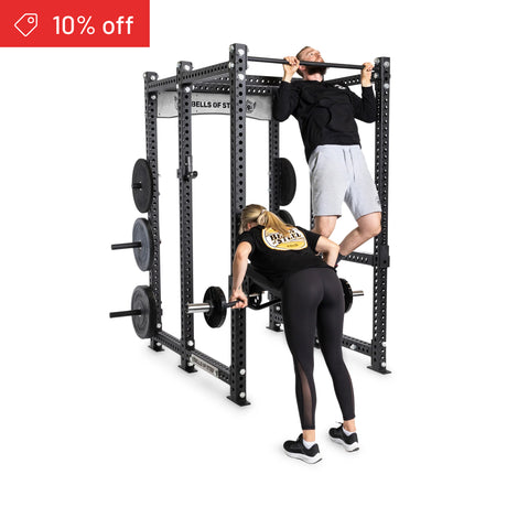 A man does a pull-up on the Bells of Steel Manticore Six Post Power Rack Builder while a woman lifts a barbell nearby. Several weight plates are stored on the rack. Both wear athletic clothing. A red banner reads "10% off.