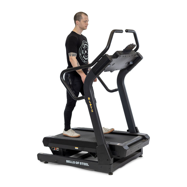 Wearing a black T-shirt and pants, a person stands on a Blitz Mountain Climber Treadmill by Bells of Steel, ready to work out on this advanced cardio machine with FTMS Bluetooth connectivity and an intuitive control panel.