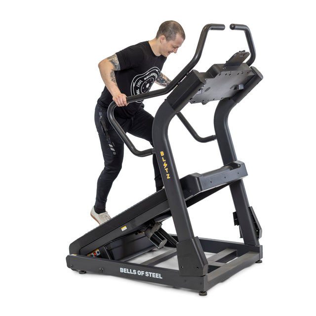 A person exercises on a Blitz Mountain Climber Treadmill, gripping the handrails while stepping in a black T-shirt and pants. This cardio machine, from Bells of Steel, offers FTMS Bluetooth connectivity for advanced tracking.