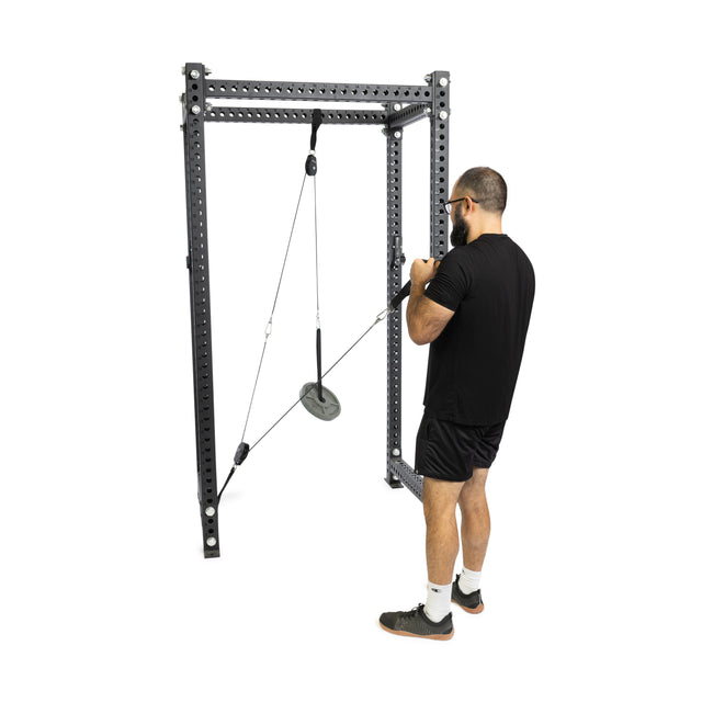 A man in a black t-shirt and shorts uses a Bells of Steel Tricep Rope Extension at the gym, performing the exercise with determination. A weight plate hangs securely by a carabiner against a plain white background.