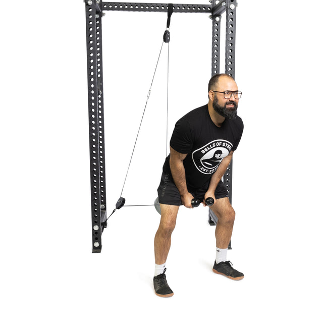 A bearded man with glasses, in a black T-shirt and shorts, precisely uses the Bells of Steel Tricep Rope Extension on a cable machine. He is bent over while working out against a crisp white background.