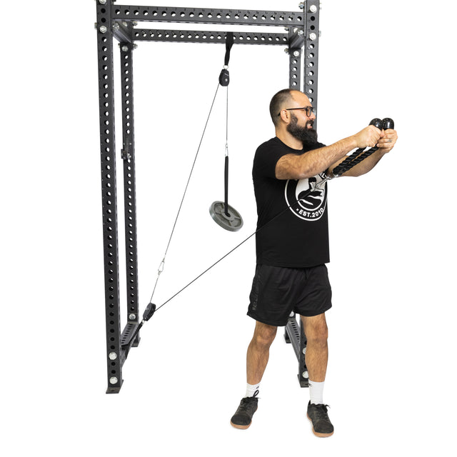 A bearded, bespectacled man in a black shirt and shorts uses the Bells of Steel Tricep Rope Extension cable machine for his workout, pulling the cables while standing in athletic shoes.