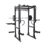 A Bells of Steel Manticore Flat Foot Power Rack (3" x 3", 1" Holes) with adjustable attachments, a barbell, weights, modular components, and safety bars is shown. The metallic frame features brand signage on top and is expertly designed for strength training exercises.