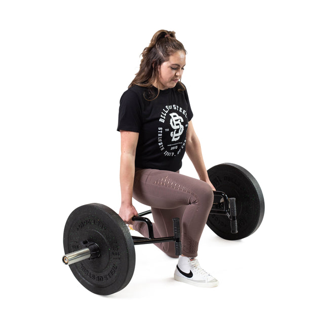 A woman in a black shirt and pink leggings kneels with one knee on the ground, gripping a Bells of Steel Open Trap Bar / Hex Bar with both hands, preparing to lift it against a white background.