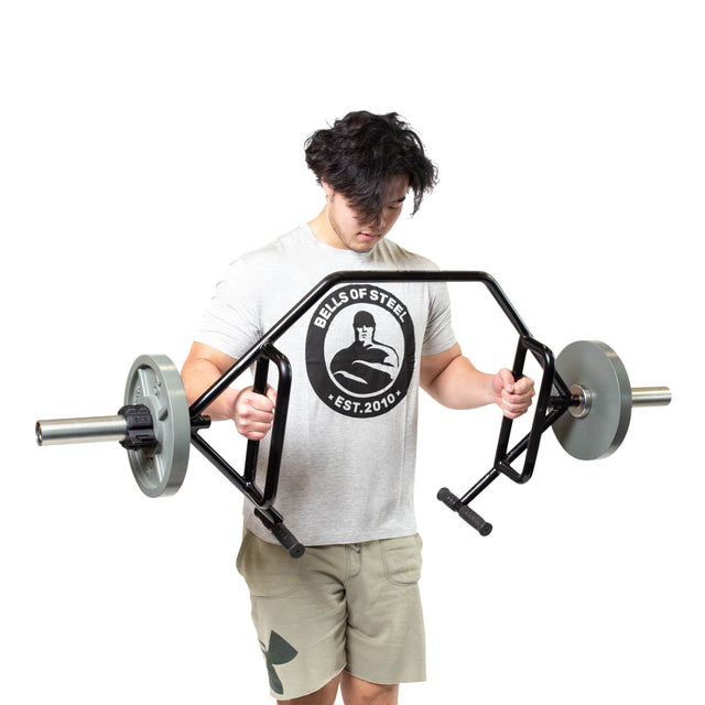 A person with dark hair holds a Bells of Steel Open Trap Bar, its rotating sleeves in action. They wear a light gray T-shirt with a logo and khaki shorts against a plain white background.