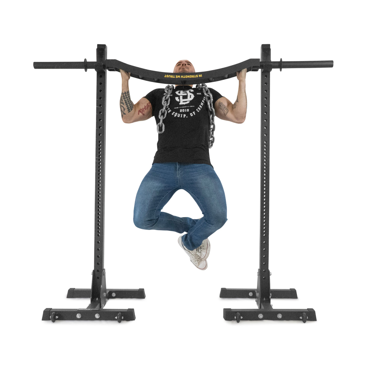 A person in a black t-shirt and jeans does a pull-up on the Bells of Steel Omni Bar, which features knurling for grip and is held by sturdy T-bar stands. Their legs are crossed at the ankles, showcasing tattoos on their arms against a plain white background.