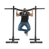 A person in a black t-shirt and jeans does a pull-up on the Bells of Steel Omni Bar, which features knurling for grip and is held by sturdy T-bar stands. Their legs are crossed at the ankles, showcasing tattoos on their arms against a plain white background.