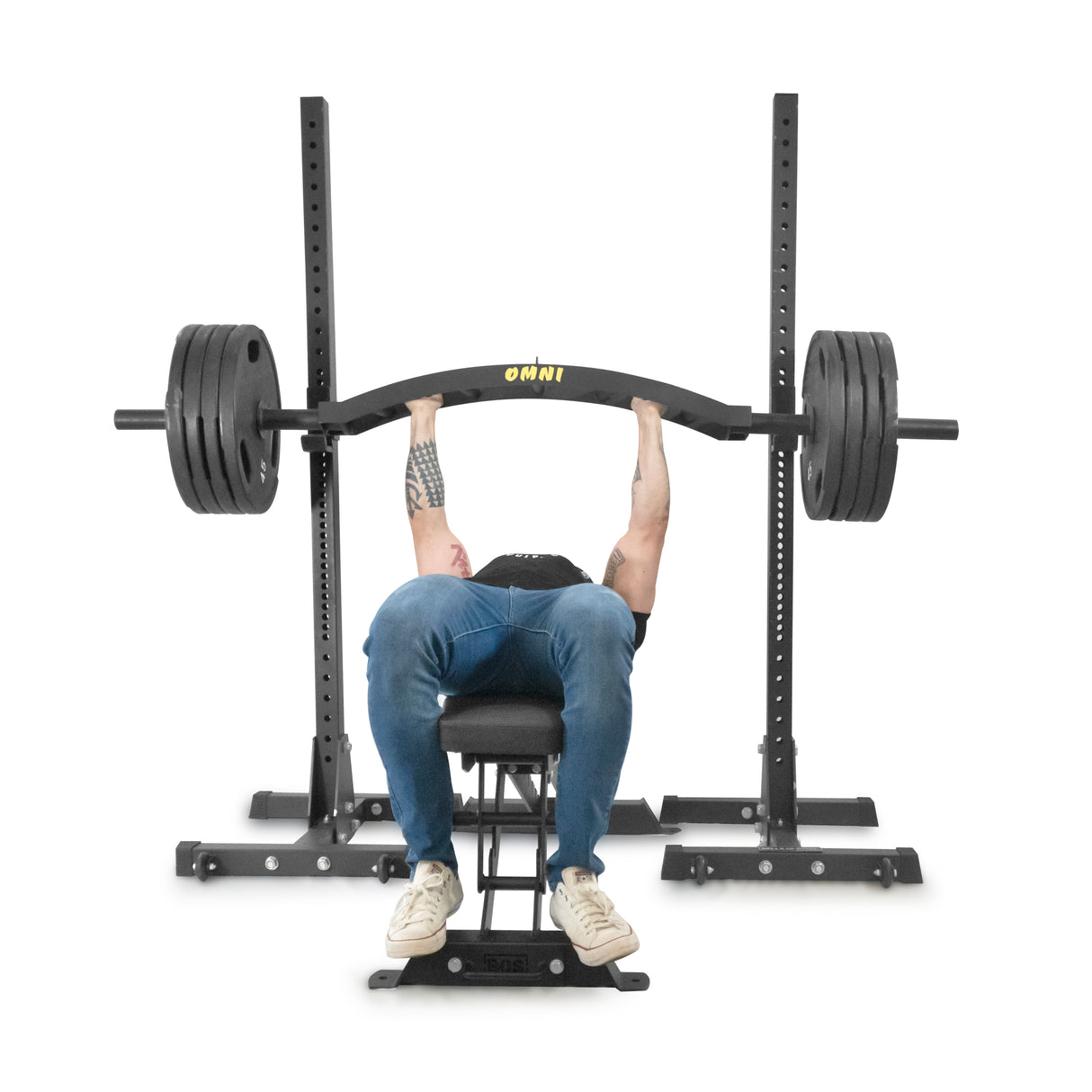 A person bench presses a heavily weighted Bells of Steel Omni Bar with distinct knurling on a sturdy power rack. They're wearing a black shirt, blue jeans, and white sneakers. The weight rack is labeled "OMNI.