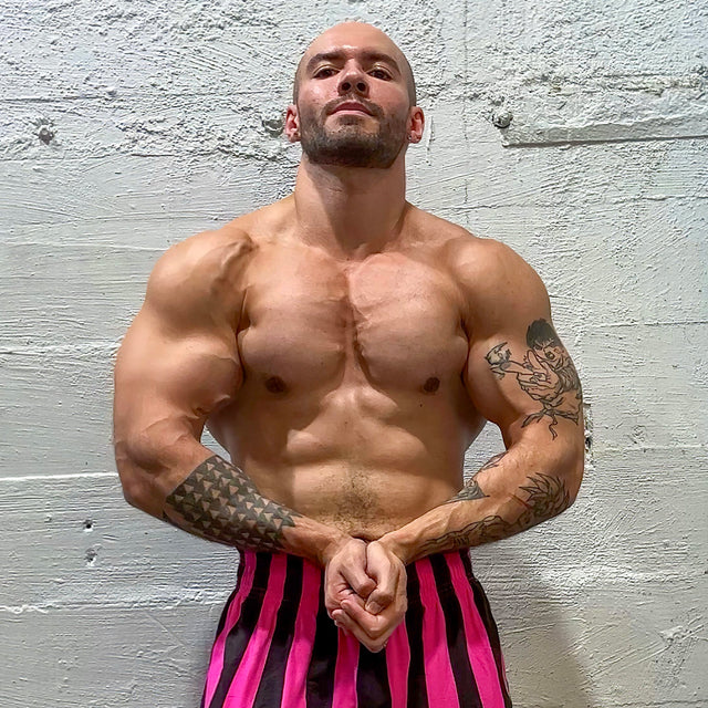 A muscular man, posing against a concrete wall in striped pink and black pants, with tattoos and clenched fists, exudes strength akin to Bells of Steel's Omni Bar with precise knurling. He's ready to conquer challenges as if in the power racks.