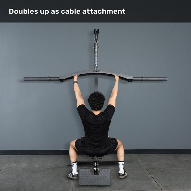 A person sits on a bench facing a gray wall, gripping the Bells of Steel Arch Nemesis Swiss Bar attached to an overhead cable machine. Text above: "Doubles up as cable attachment.