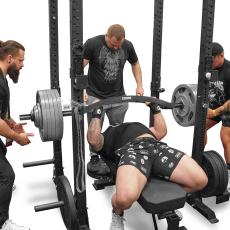 Using the Arch Nemesis Swiss Bar by Bells of Steel, a person performs a bench press on a power rack with three spotters' assistance. The lifter, wearing a black shirt and patterned shorts, presses the bar with angled grips while two spotters stand behind and one stands to the side.