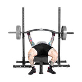 A tattooed individual in a gray shirt, black shorts, and weightlifting gloves lies on a bench press using the Bells of Steel Arch Nemesis Swiss Bar with angled grips. The sturdy gym equipment is black.