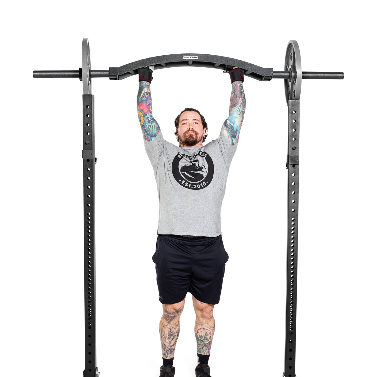 A tattooed person does a pull-up with angled grips on a Bells of Steel Arch Nemesis Swiss Bar, attached to a power rack. Wearing a gray t-shirt, black shorts, and gloves, they stand out against the plain white background.