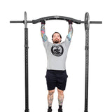 A tattooed person does a pull-up with angled grips on a Bells of Steel Arch Nemesis Swiss Bar, attached to a power rack. Wearing a gray t-shirt, black shorts, and gloves, they stand out against the plain white background.